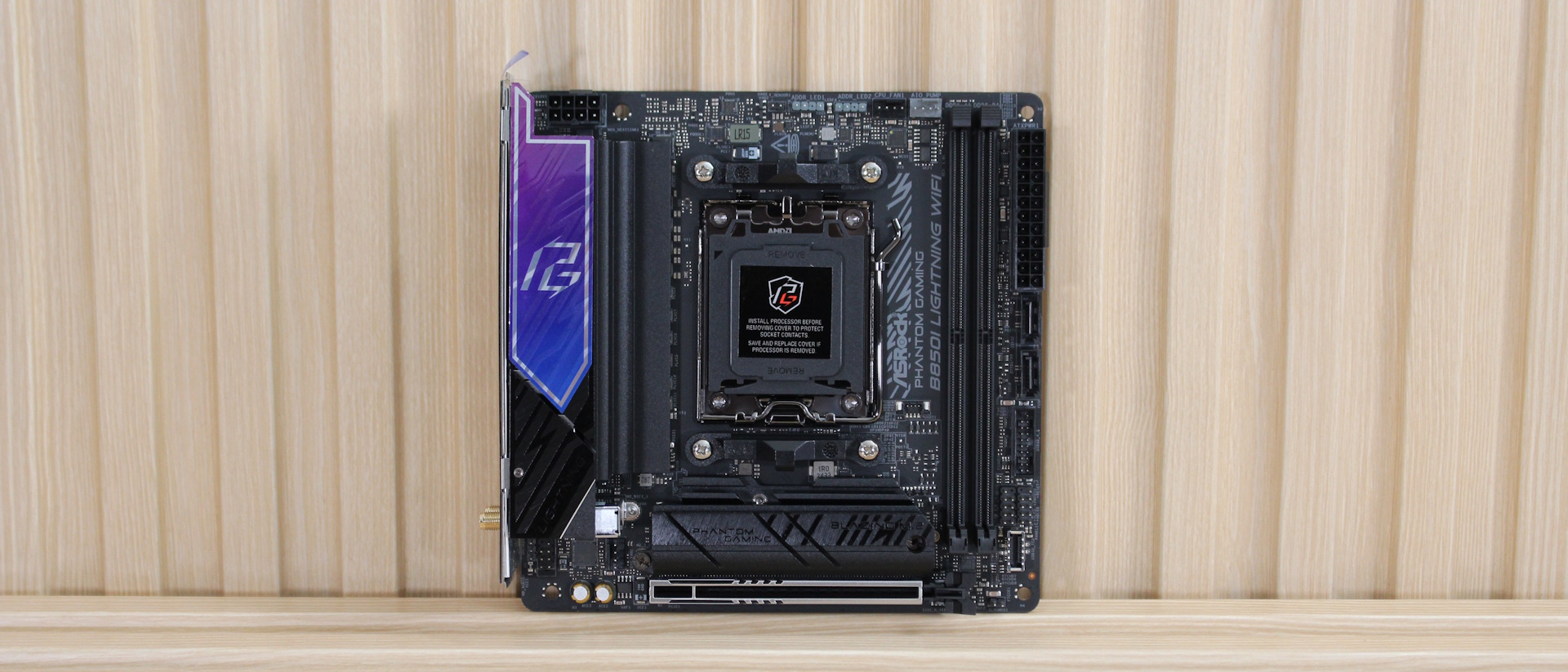 Материнская плата ASRock B850I Lightning Mini-ITX, прислоненная к решетчатой стене. Эта материнская плата не включает встроенную RGB-подсветку, но металлический логотип PG (phantom gaming) над IO-обшивкой, который плавно переходит от фиолетового к синему, добавляет визуального стиля.